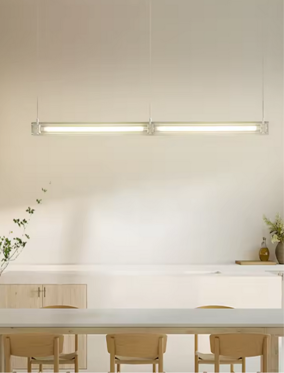 A modern, sleek linear glass pendant light with stainless steel accents, suspended over a counter, dispersing a warm glow.