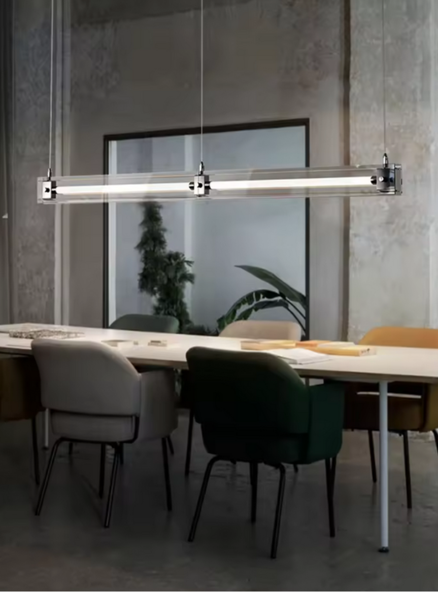 A modern, sleek linear glass pendant light with stainless steel accents, suspended over a counter, dispersing a warm glow.