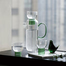 tall round clear glass pitcher with green handle and cup with green base